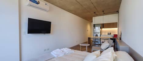 A modern hotel room with a bed, a flat-screen TV, a small round table, and a kitchenette in the background.