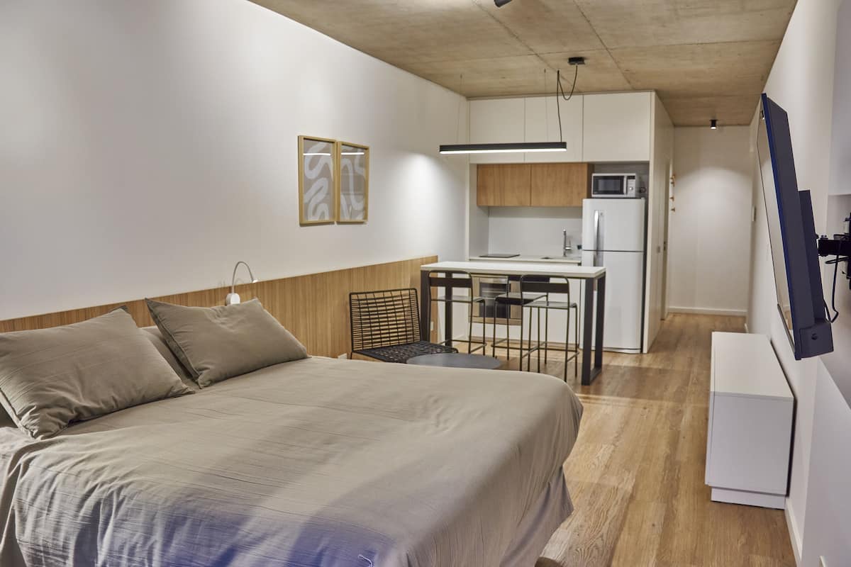 A compact hotel room with a bed, a small kitchenette, a dining area, and a TV mounted on the wall.
