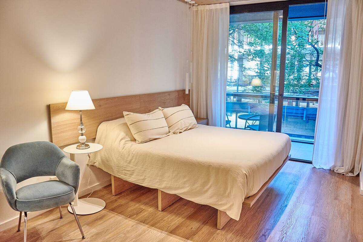 A hotel room with a bed, a chair, a lamp, and a view of an outdoor area.