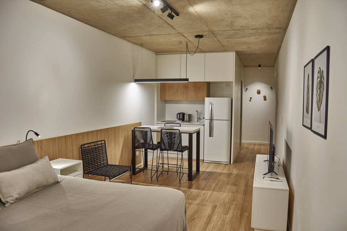 A compact hotel room with a bed, a small dining table with chairs, a kitchenette, and a desk with a computer.
