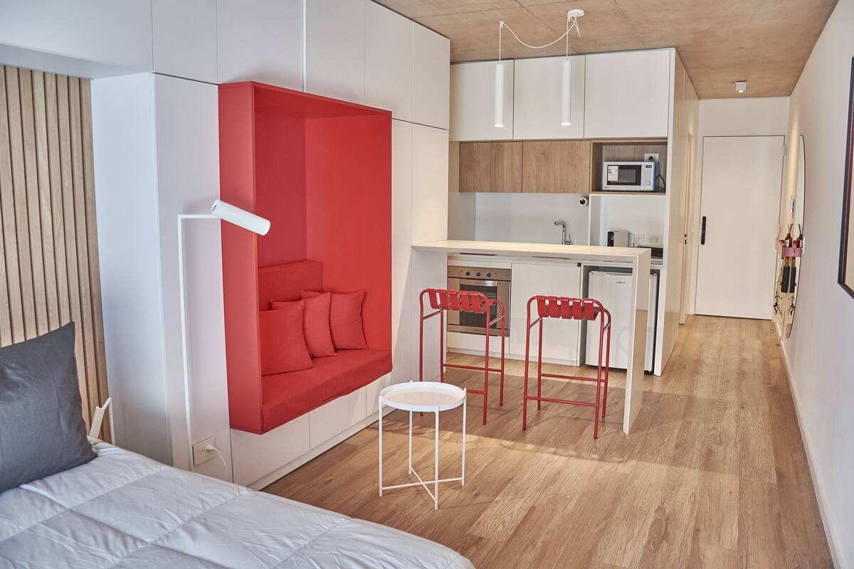 A compact living space with a kitchenette, a red accent wall, a bed, and a small white table.