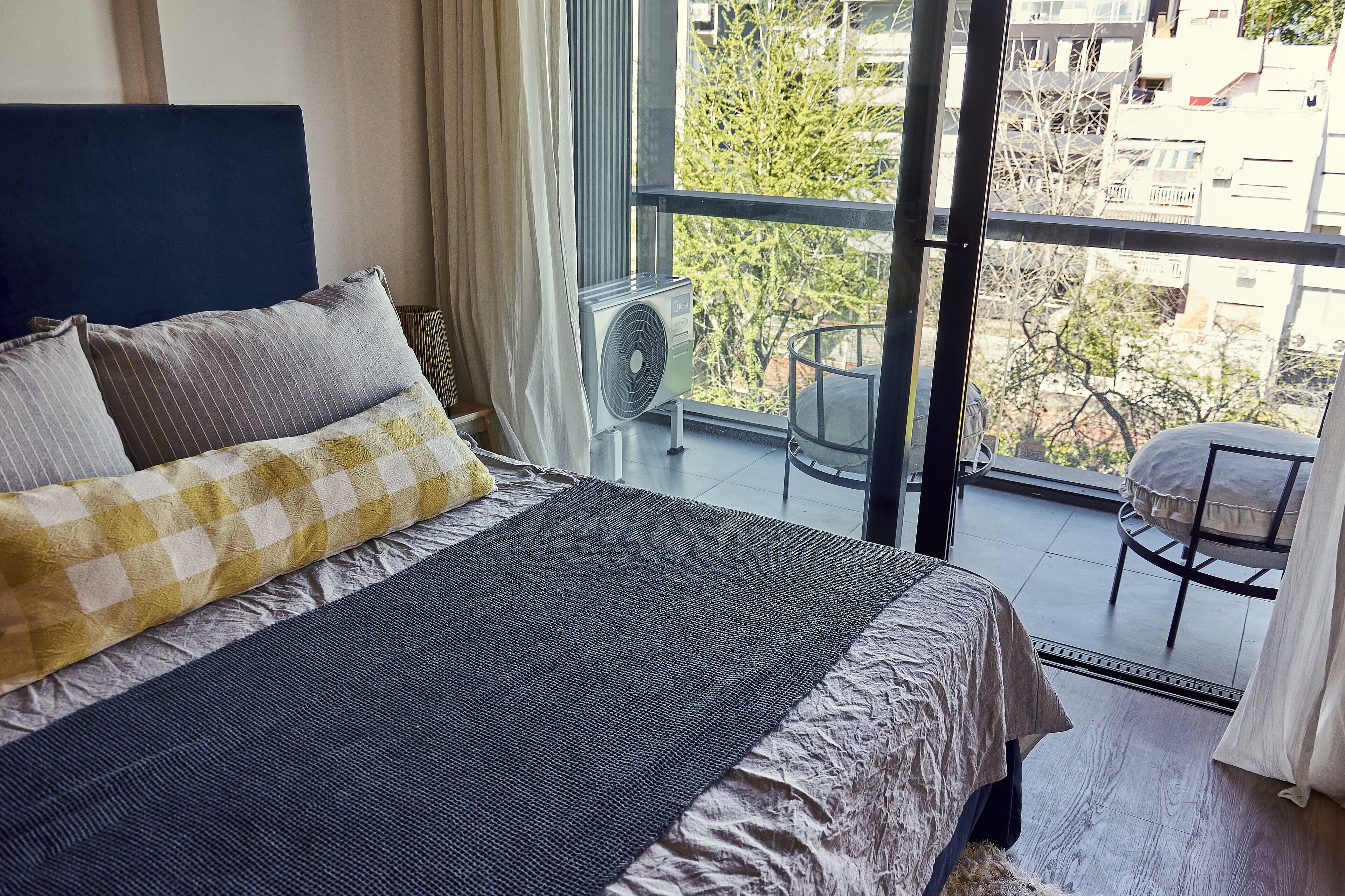 Un dormitorio con una cama grande, una ventana con vistas a edificios y árboles, y un balcón con una silla y una mesa.