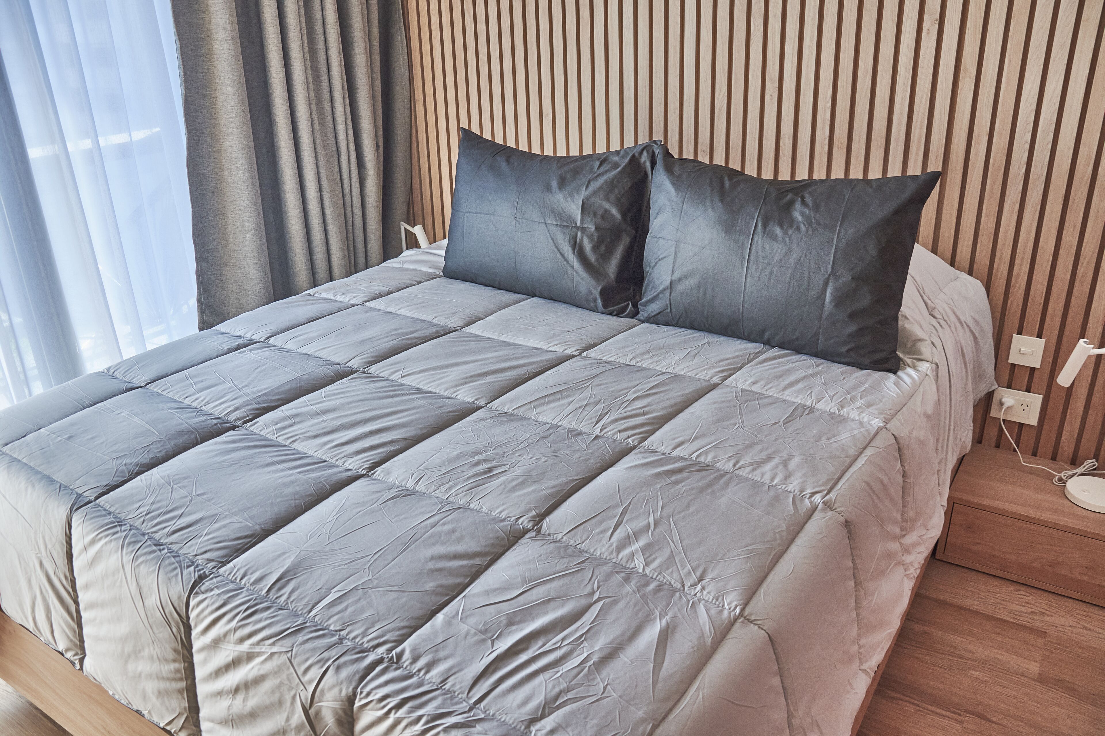 Una cama bien hecha con colcha gris y dos almohadas, cabecera de madera y mesita de noche con toma de corriente.