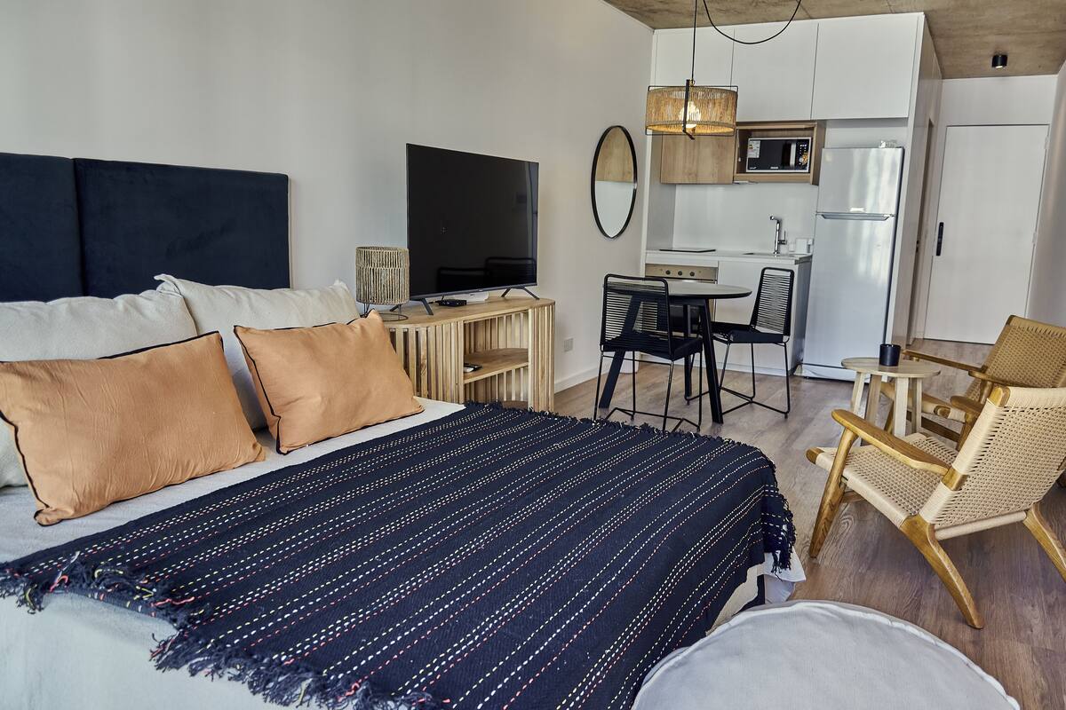 A bedroom with a large bed, a TV, a small kitchenette, and a dining area.