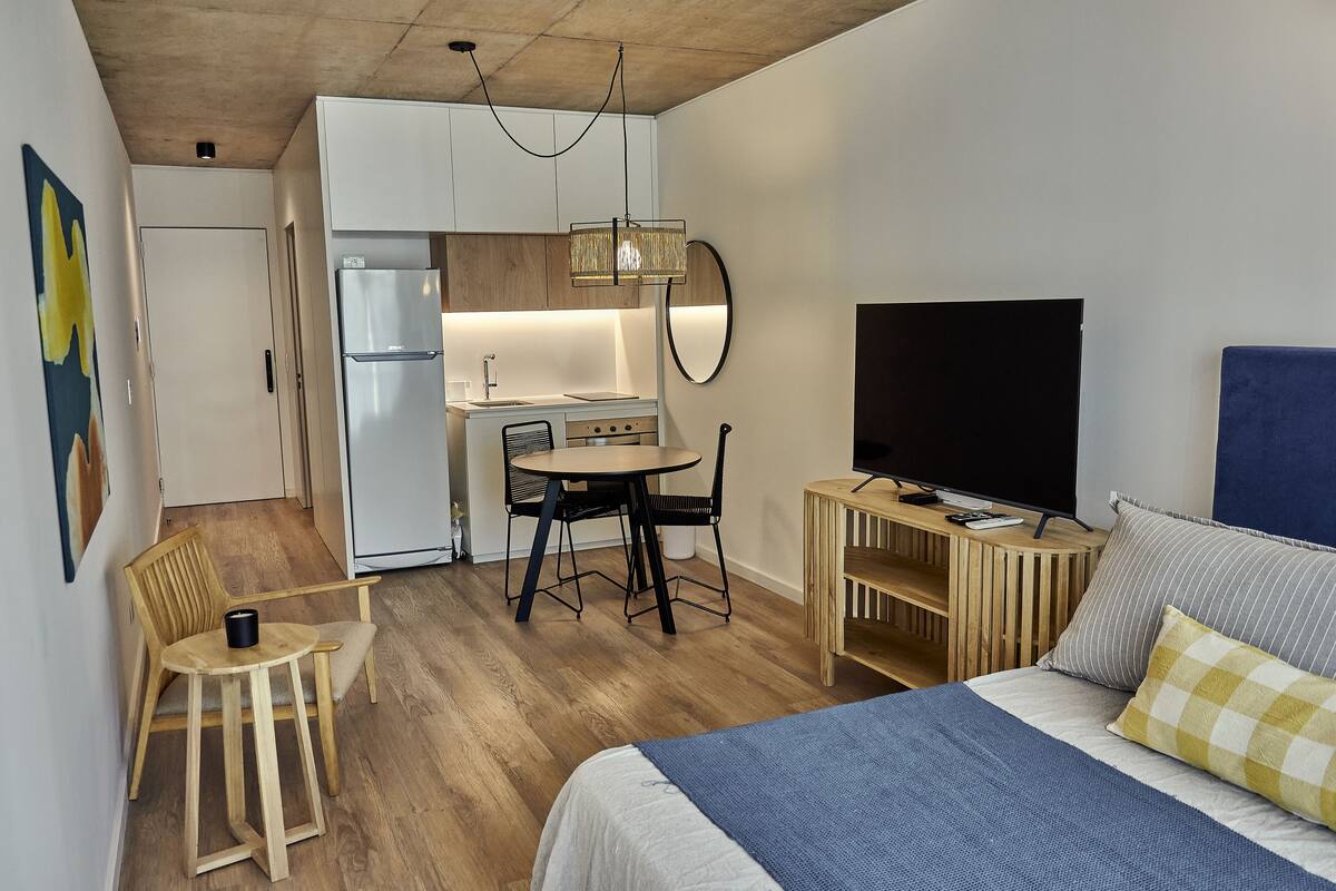 A compact hotel room with a bed, a desk with a TV, a small kitchenette, and a dining area.