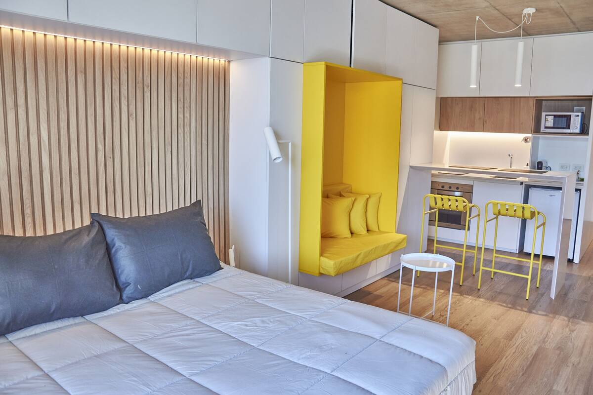A modern bedroom with a large bed, a built-in desk, and a kitchen area with yellow chairs.