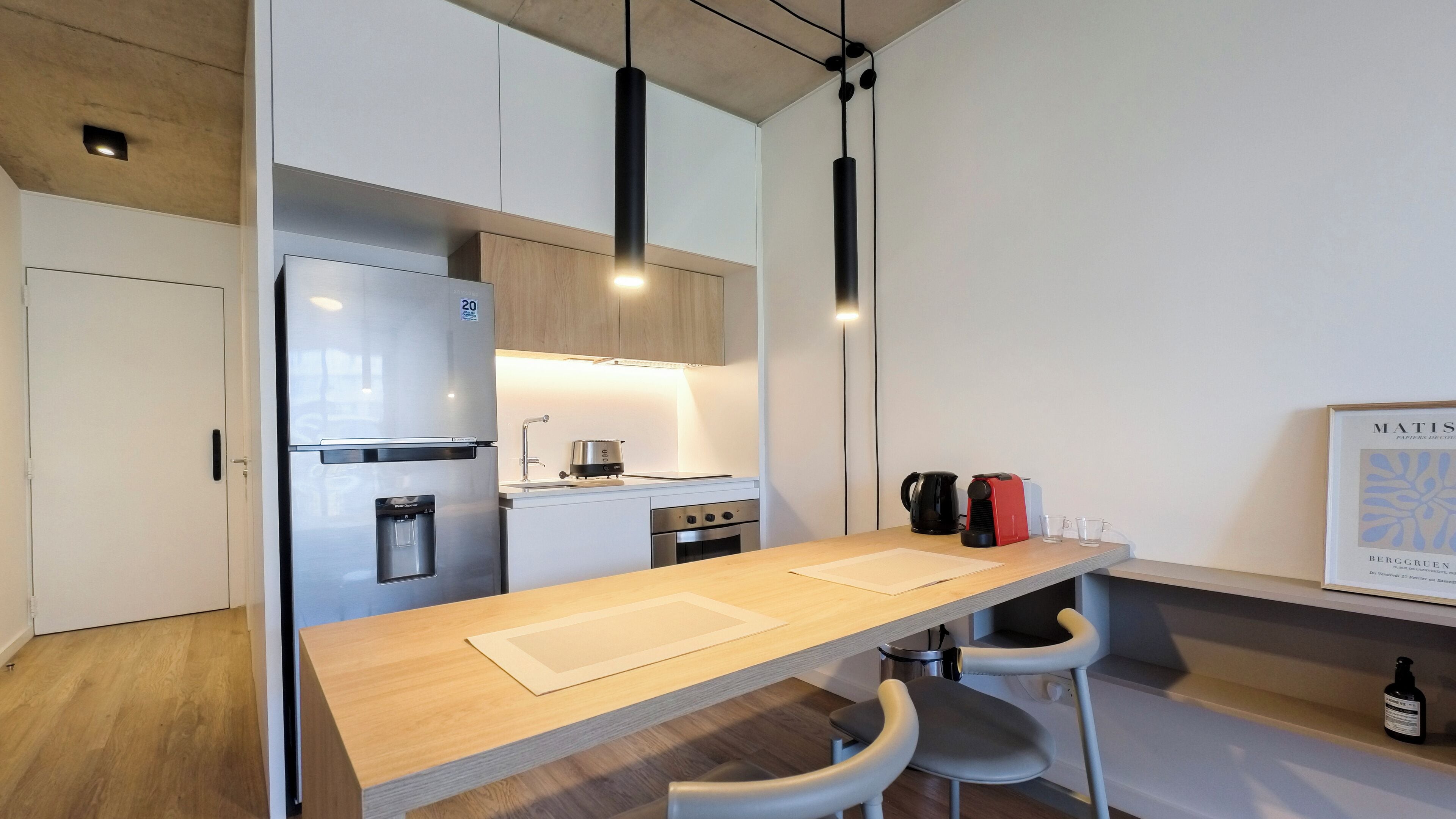 A modern kitchen with a stainless steel refrigerator, a built-in water dispenser, a stove, and a coffee maker. There are two white chairs and a wooden table.