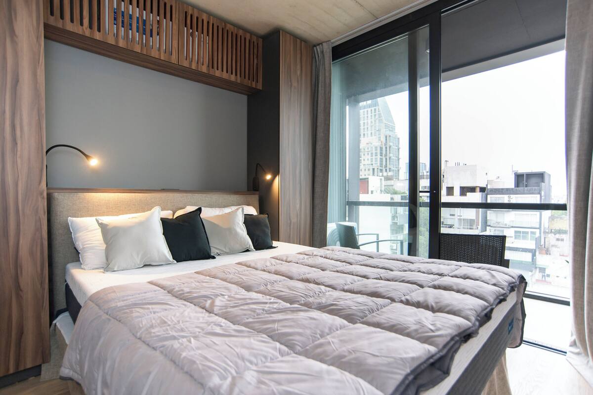 A modern hotel room with a large bed, a wooden headboard, and a view of the city through floor-to-ceiling windows.