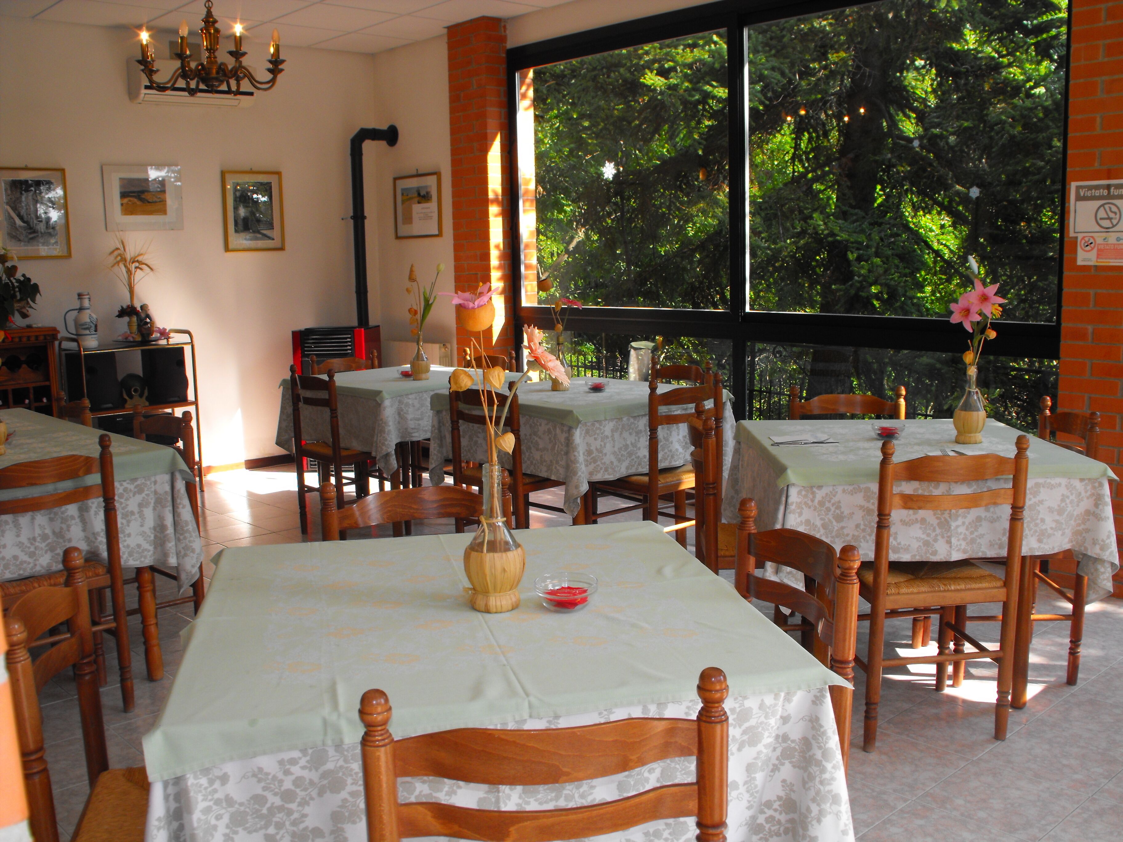 A dining area with tables and chairs, a chandelier, and a view of greenery through large windows.