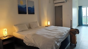 A hotel room with a bed, bedside lamps, a suitcase, an air conditioning unit, and a door. - Hotel Auro Maison (Puducherry)