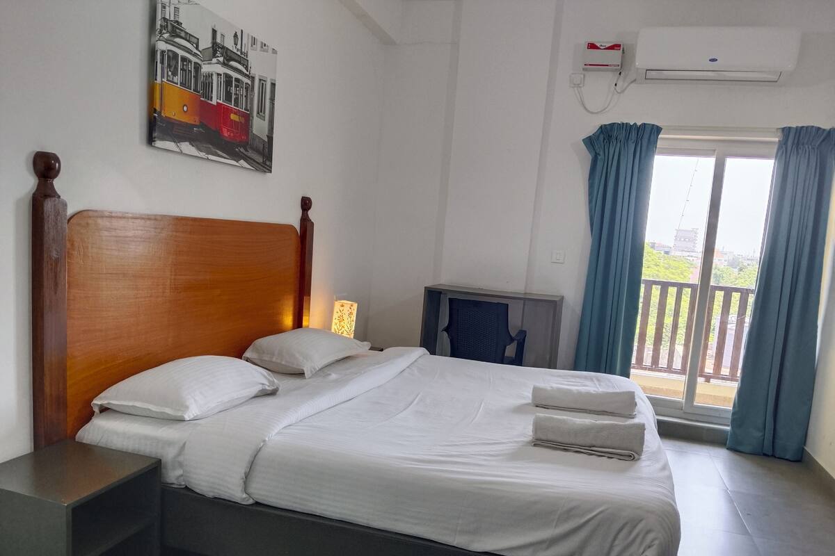 A hotel room with a wooden headboard bed, a nightstand, a desk, a chair, a window with curtains, and an air conditioning unit.