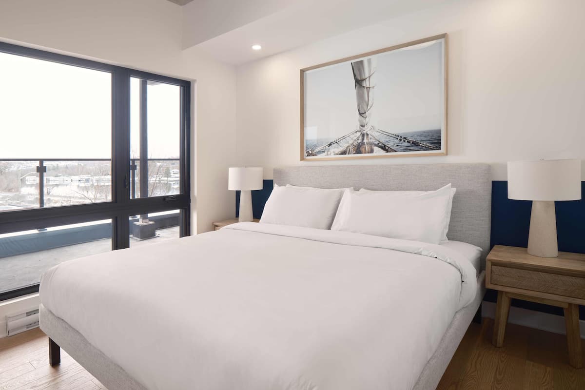 A modern hotel room with a large bed, a view through a window, and a framed artwork above the bed.
