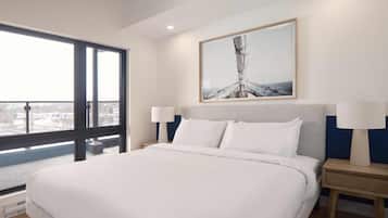 A modern hotel room with a large bed, a view through a window, and a framed artwork above the bed.