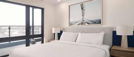 A modern hotel room with a large bed, a view through a window, and a framed artwork above the bed.