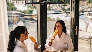 Two people in white robes at a round table with breakfast items, a view of a marina, and a chandelier. - Quartier des Marinas - Hôtel Expérience (Magog)