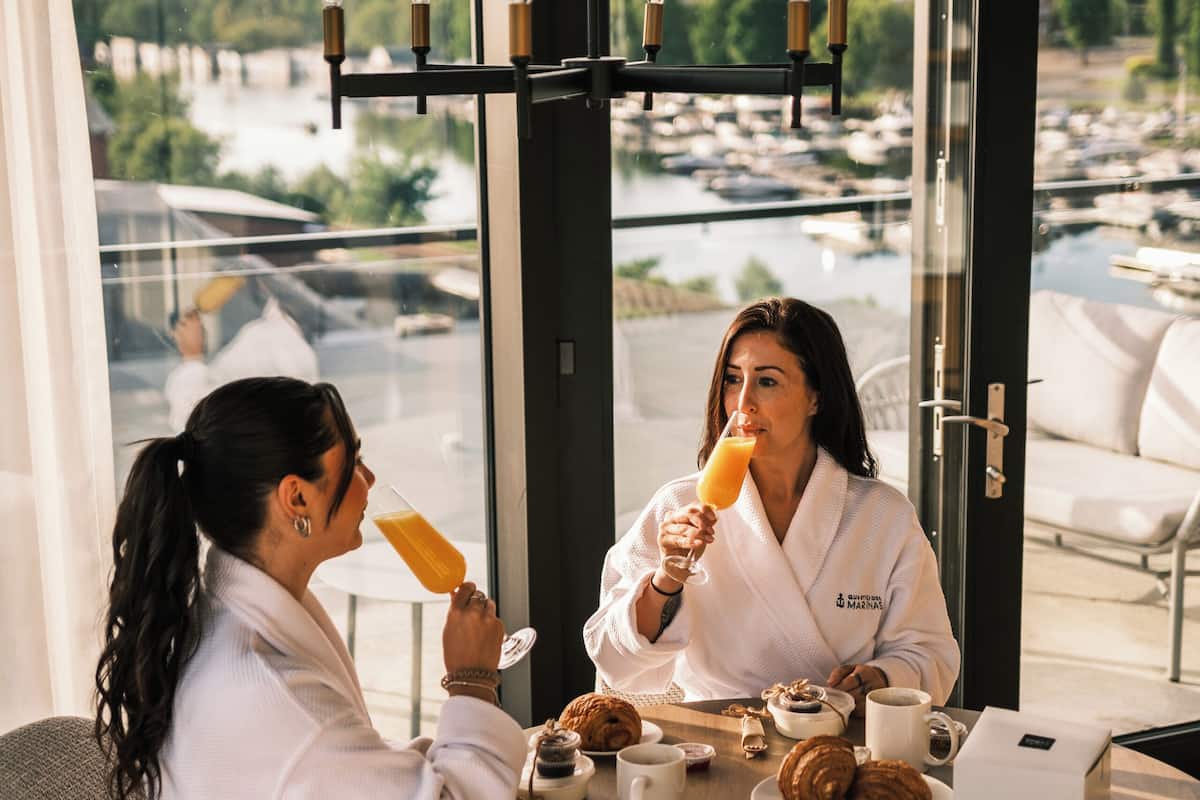 Two people in white robes at a round table with breakfast items, a view of a marina, and a chandelier.