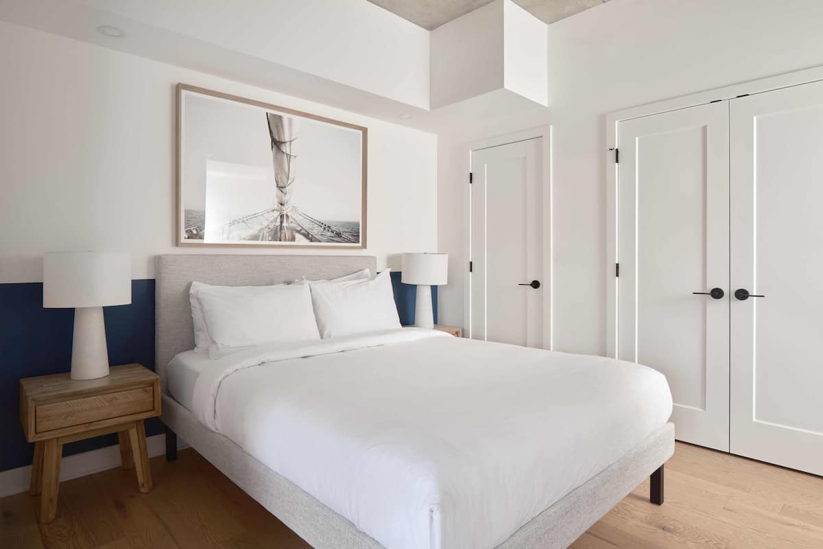 A modern bedroom with a large bed, two bedside tables with lamps, a framed artwork above the bed, and a set of white doors.