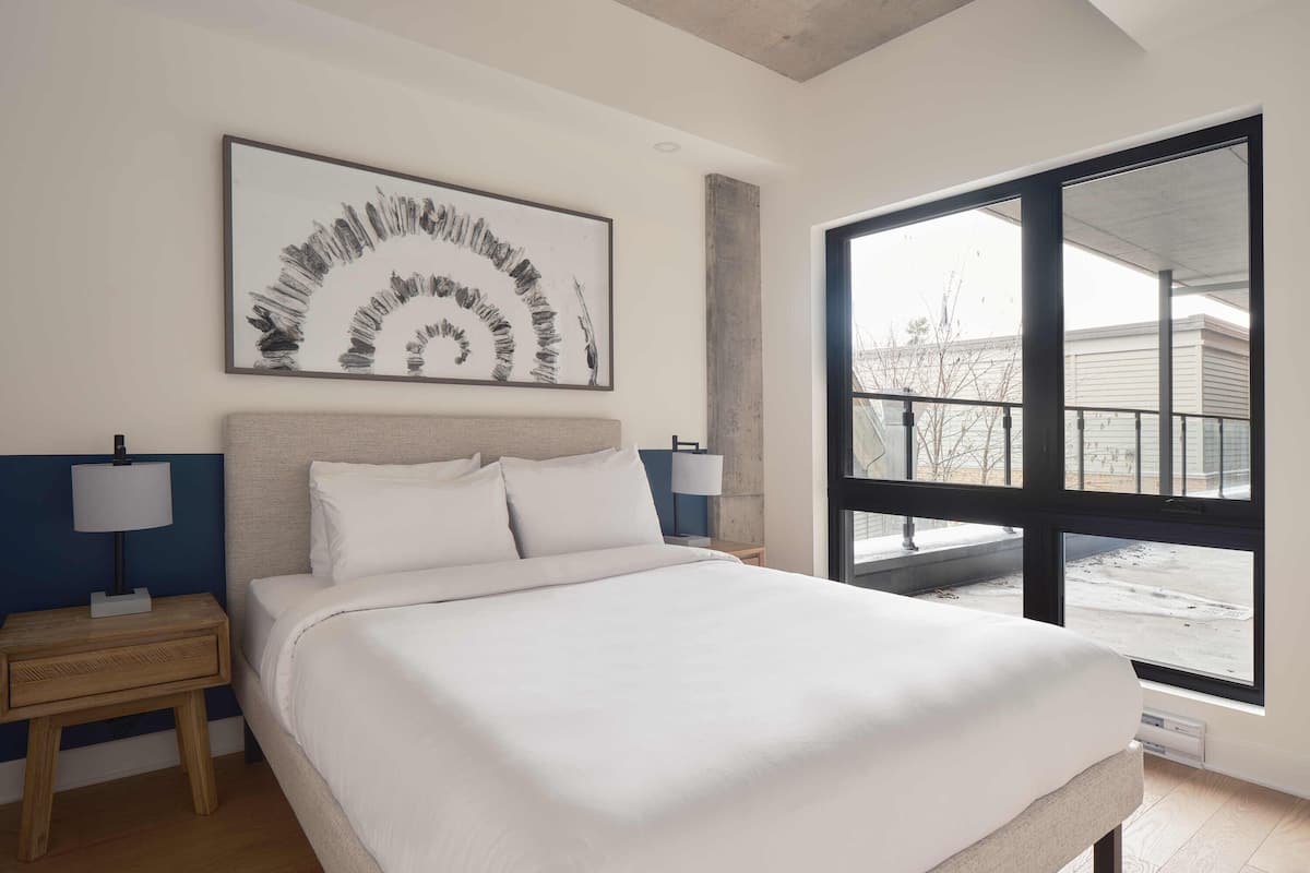 A bedroom with a large bed, a nightstand, two bedside lamps, a large window with a view of the outside, and a modern abstract painting on the wall.