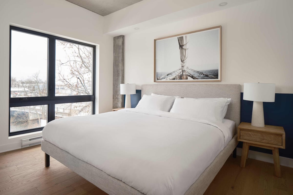 A modern hotel room with a large bed, two bedside tables, a large window, and a framed artwork on the wall.