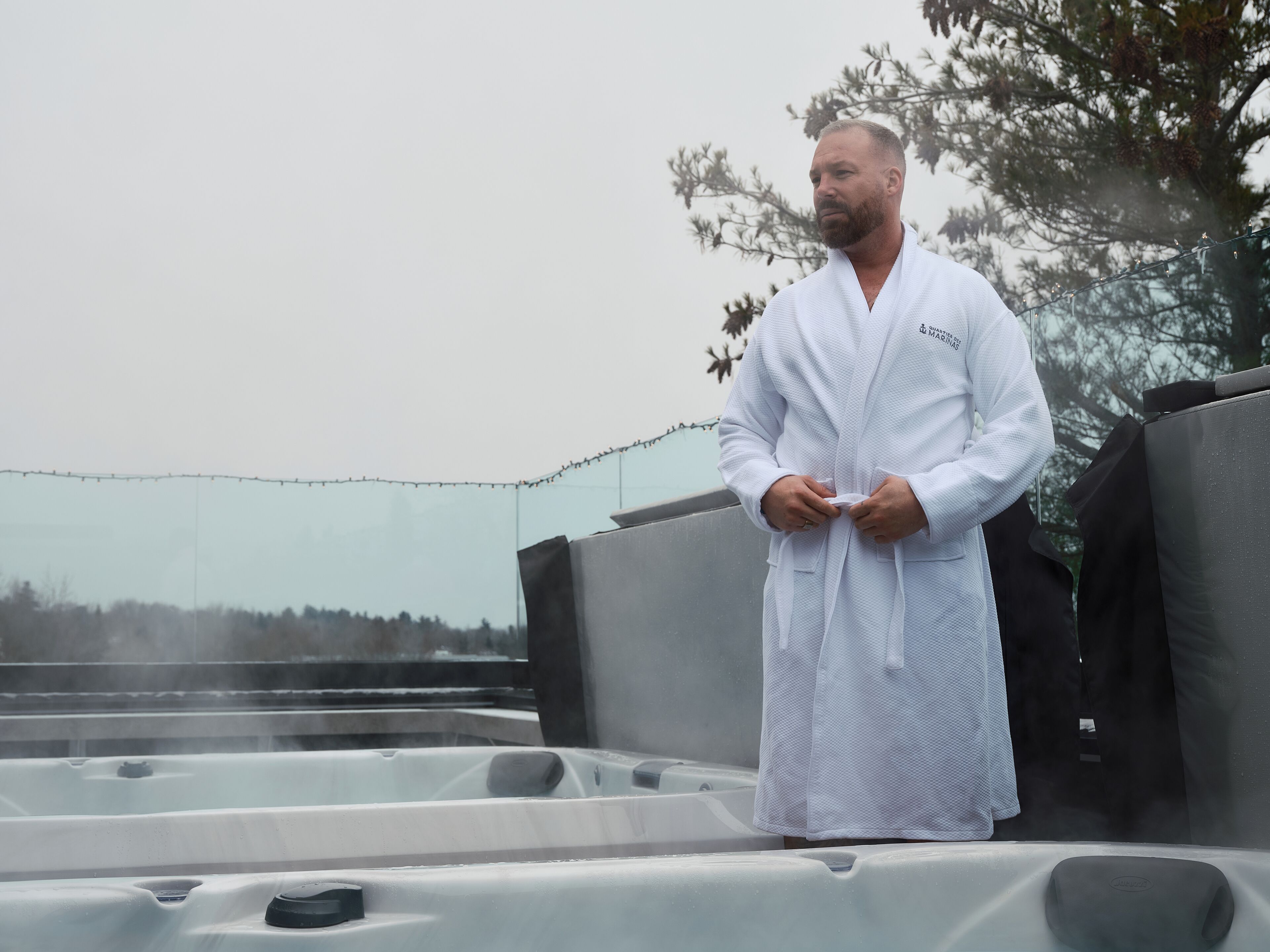 Une personne vêtue d’un peignoir blanc se tient près d’un jacuzzi dont s’échappe de la vapeur, entourée d’arbres et d’une guirlande lumineuse.
