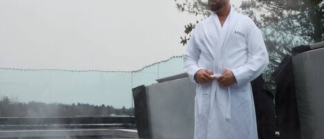 Une personne vêtue d’un peignoir blanc se tient près d’un jacuzzi dont s’échappe de la vapeur, entourée d’arbres et d’une guirlande lumineuse.