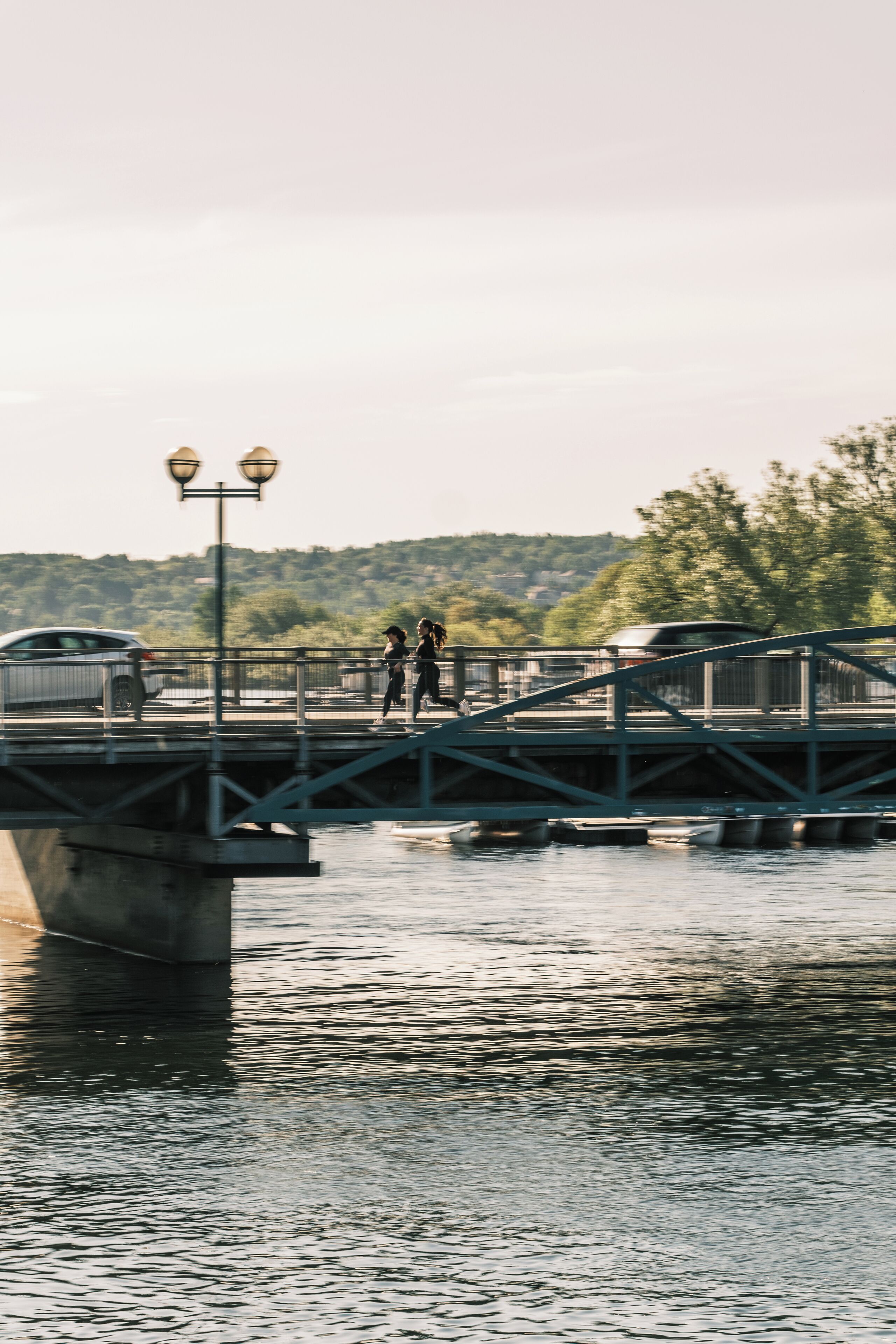 Un pont piétonnier enjambant une étendue d’eau, sur lequel se trouvent deux personnes qui marchent, une voiture et un lampadaire.