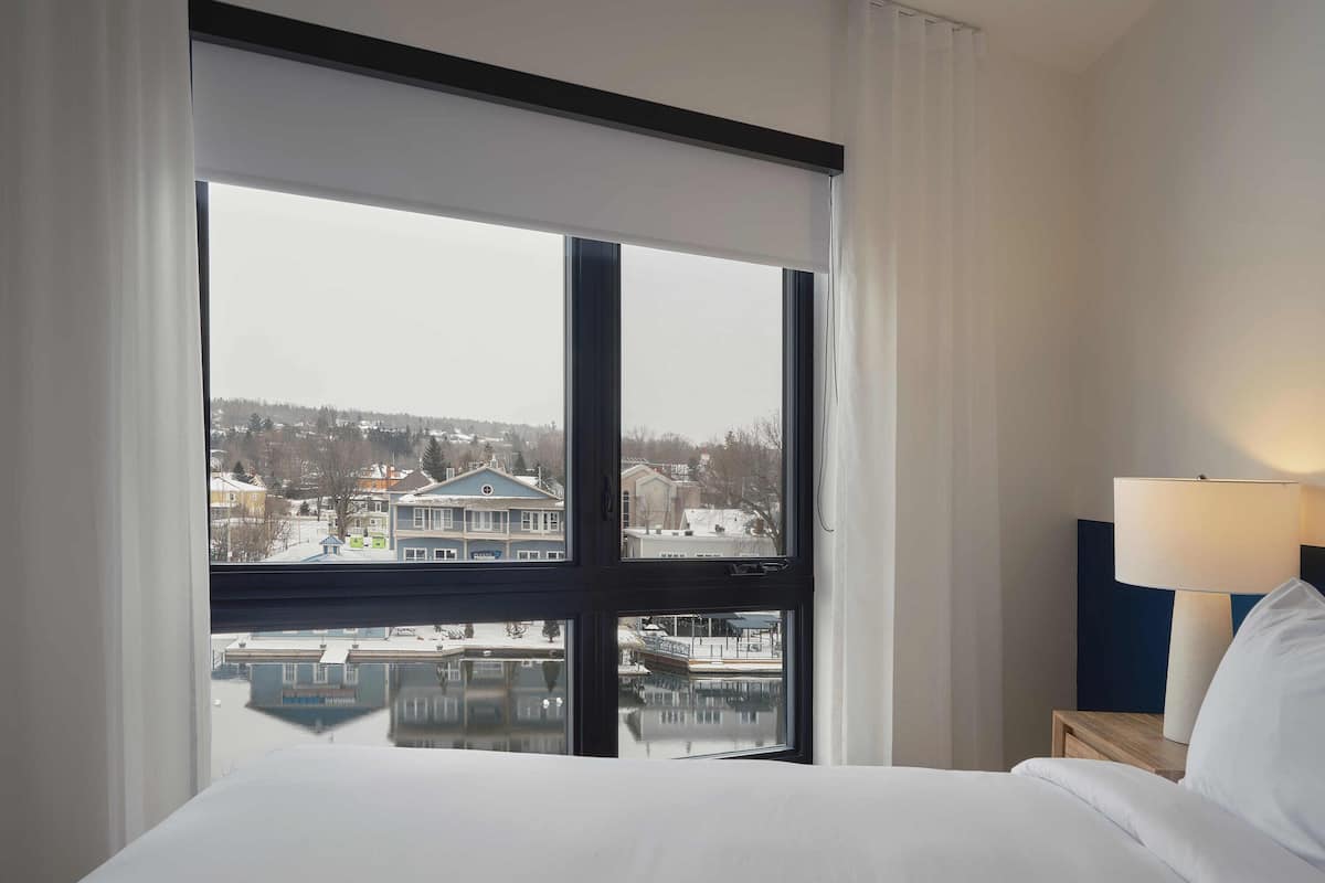 A hotel room with a large window, a bed, a nightstand with a lamp, and a view of buildings and water outside.