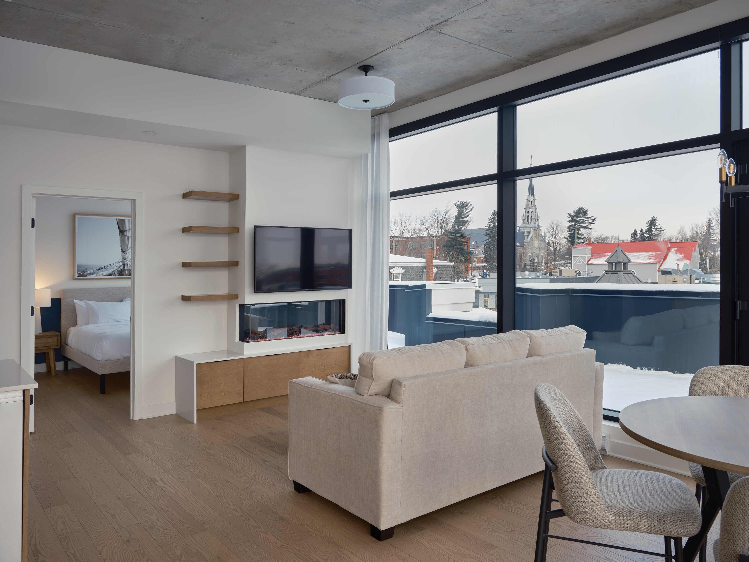 A modern hotel room with a large window, a flat-screen TV, a sofa, and a dining area.