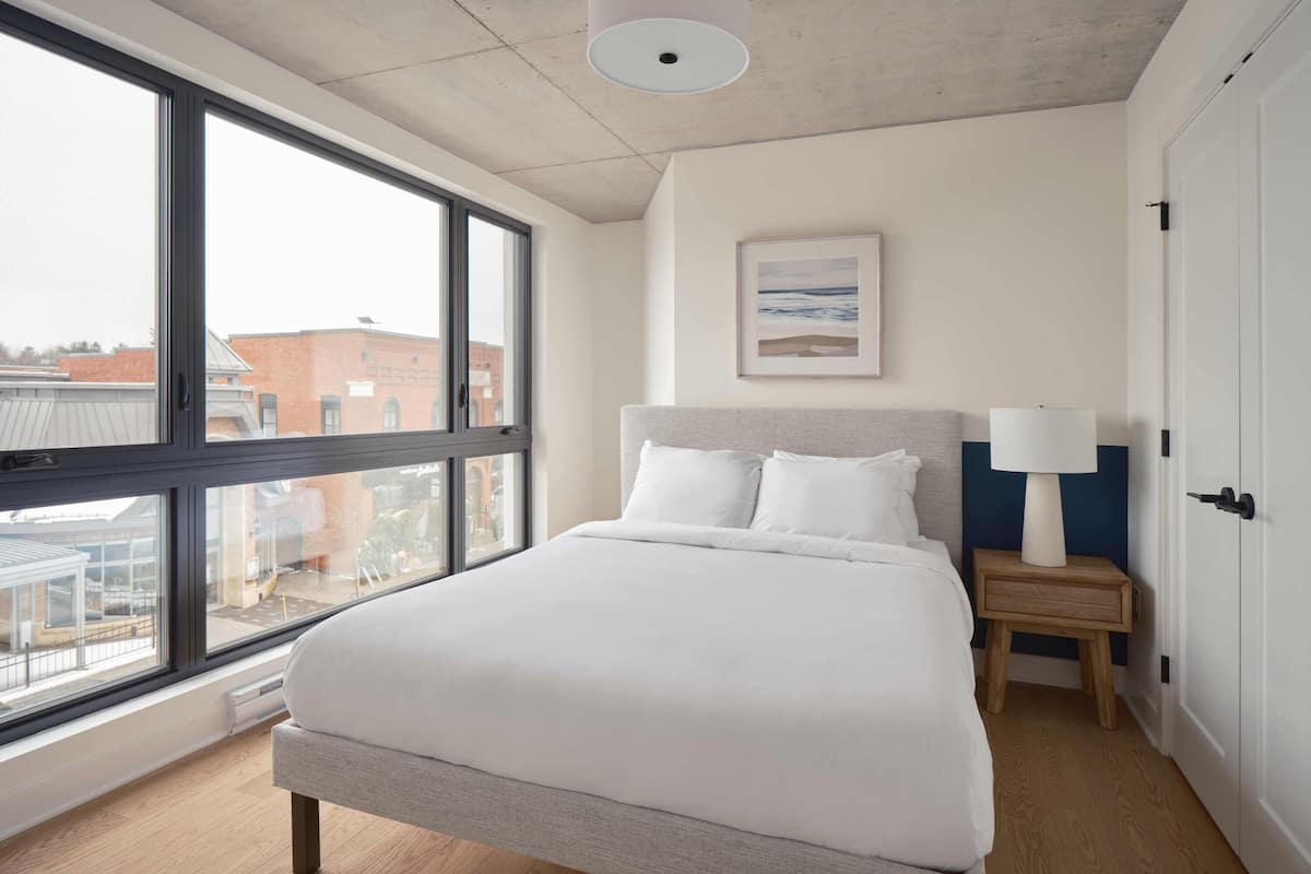 A modern hotel room with a large bed, a bedside table with a lamp, a framed picture on the wall, and a view of buildings through large windows.