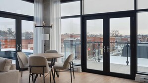 A modern dining area with a round table and chairs, large windows with a city view, and a pendant light. - Quartier des Marinas - Hôtel Expérience (Magog)