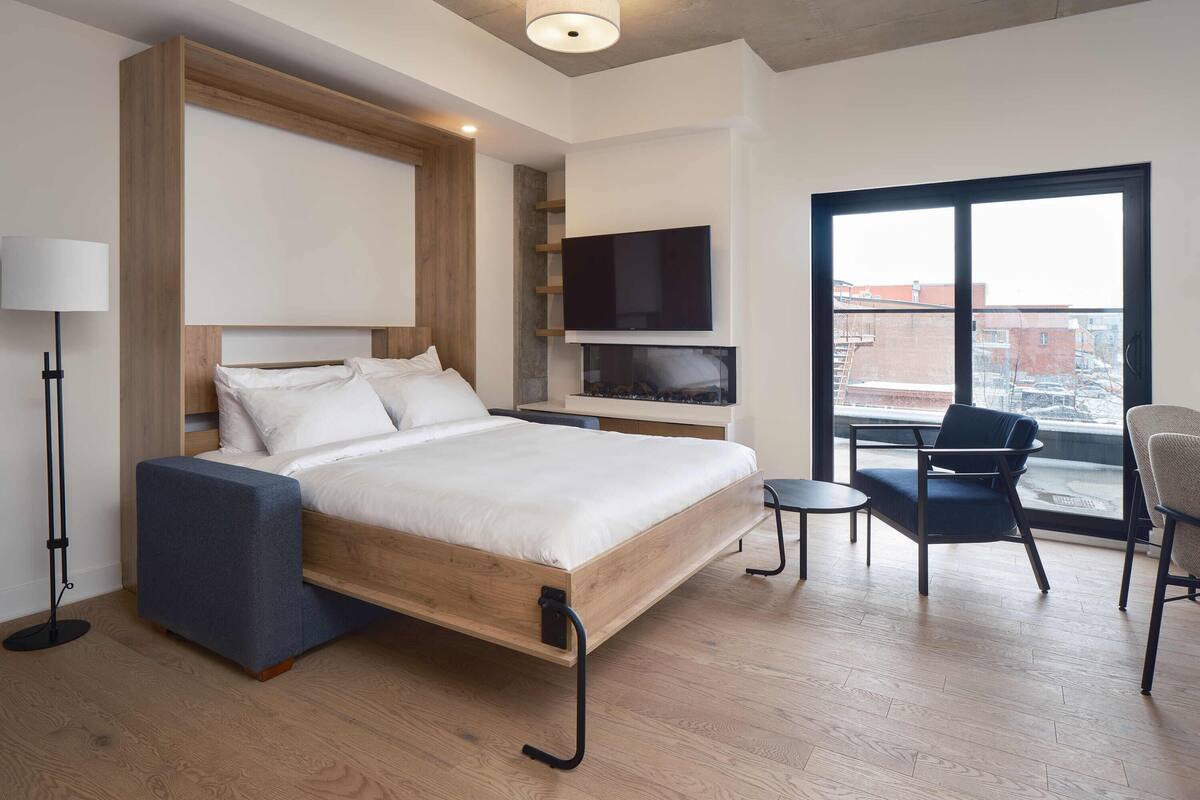 A modern hotel room with a wooden bed, a flat-screen TV, a small table, and a chair.