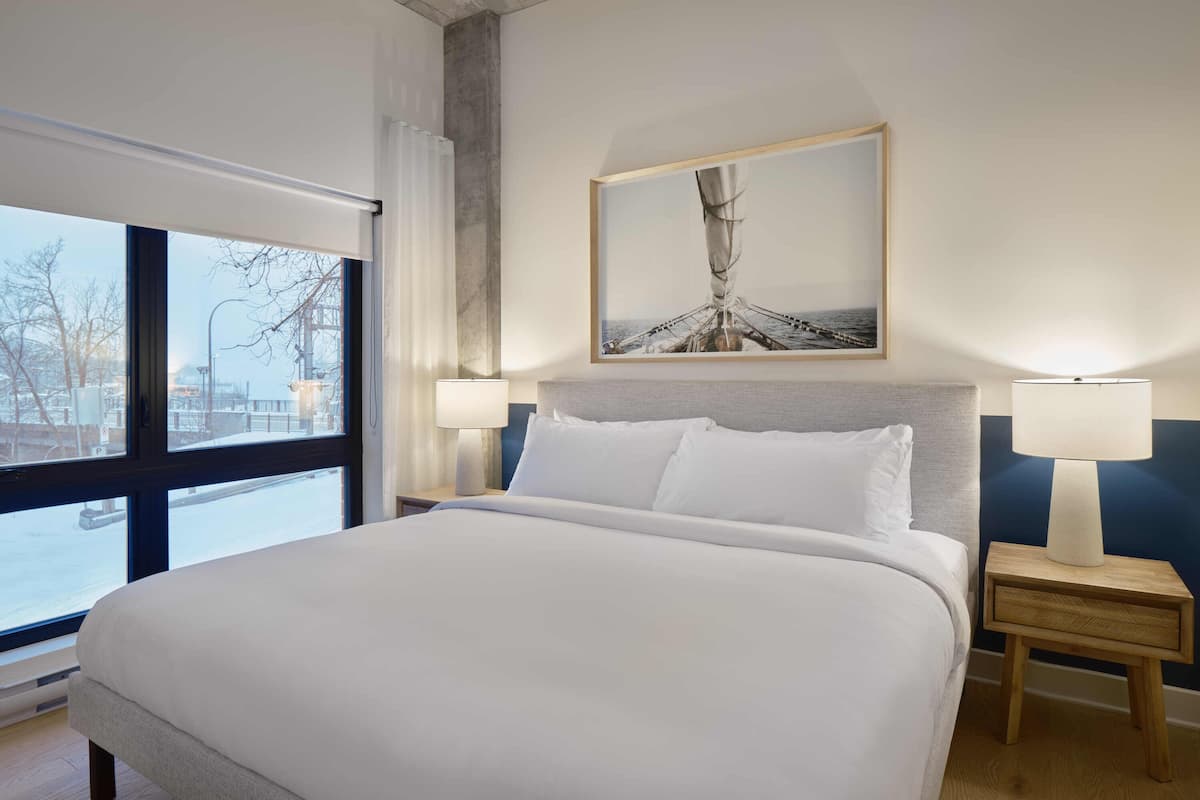 A large bed with white linens, two bedside tables with lamps, a painting above the bed, and a view of a snowy street through a large window.