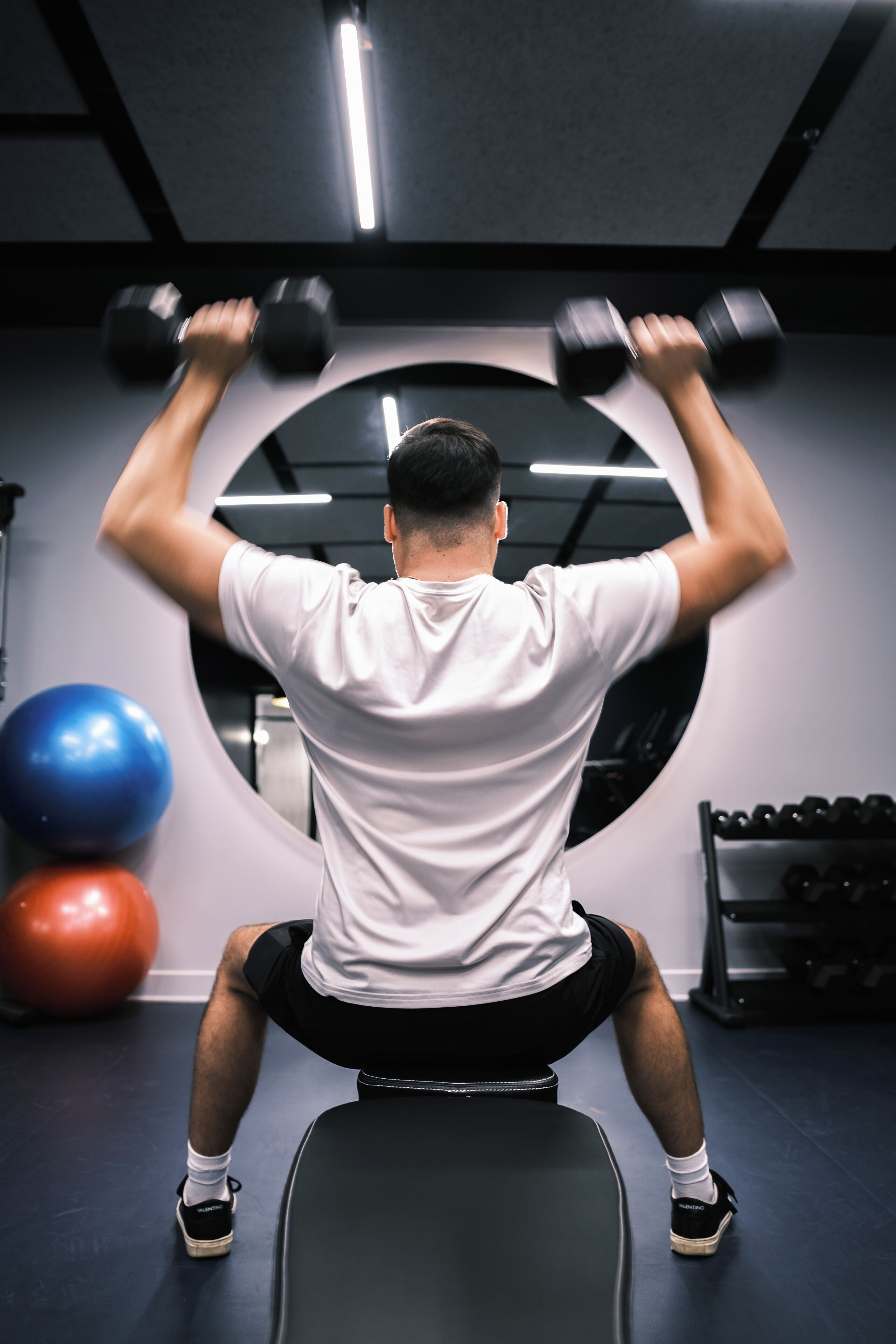 A person performing a seated dumbbell exercise in a gym with exercise balls and a bench.