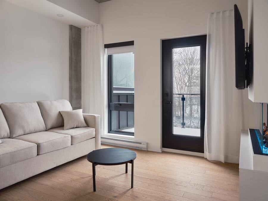 Una sala de estar moderna con un sofá, una mesita redonda y un ventanal con vistas a árboles.
