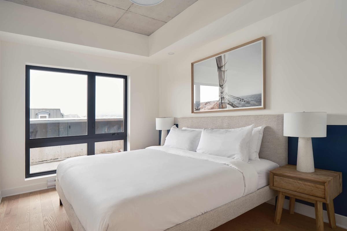 A hotel room with a large bed, two bedside lamps, a wooden nightstand, a large window with a view, and a framed picture on the wall.