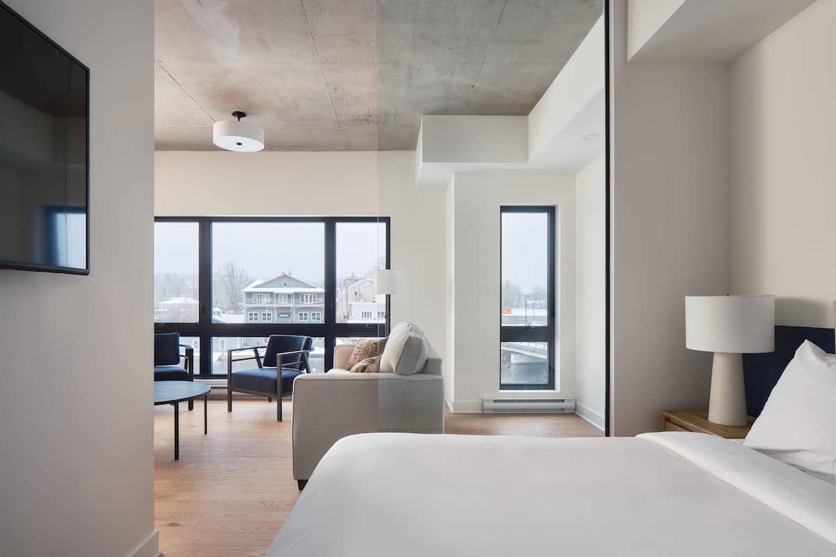 A modern hotel room with a large bed, a desk, a chair, and a view of a cityscape through large windows.