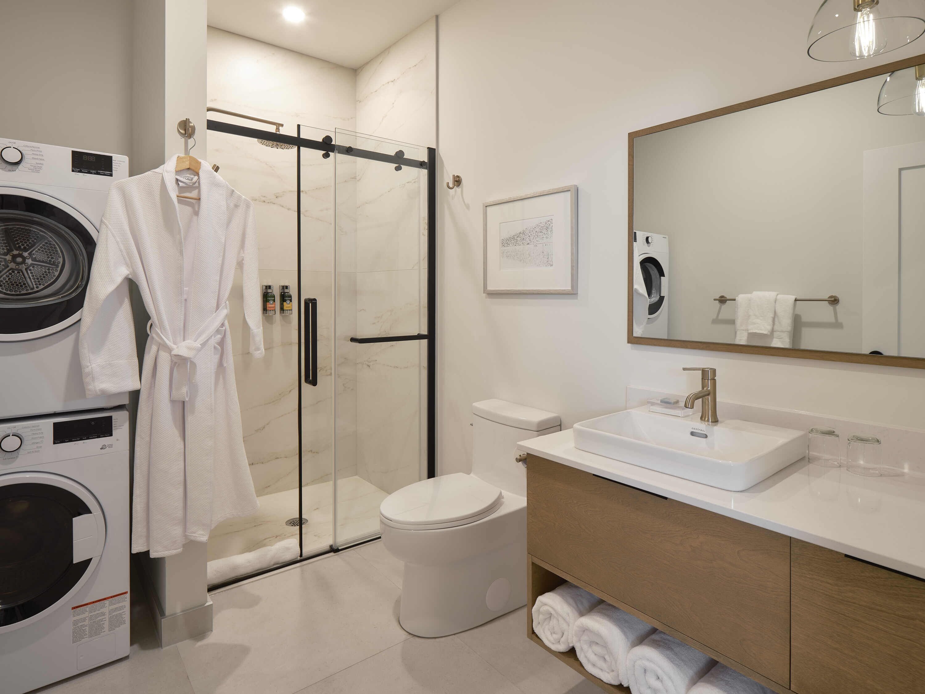 Bany modern amb boxa de dutxa de vidre, rentadora integrada, vàter i un lavabo amb mirall.