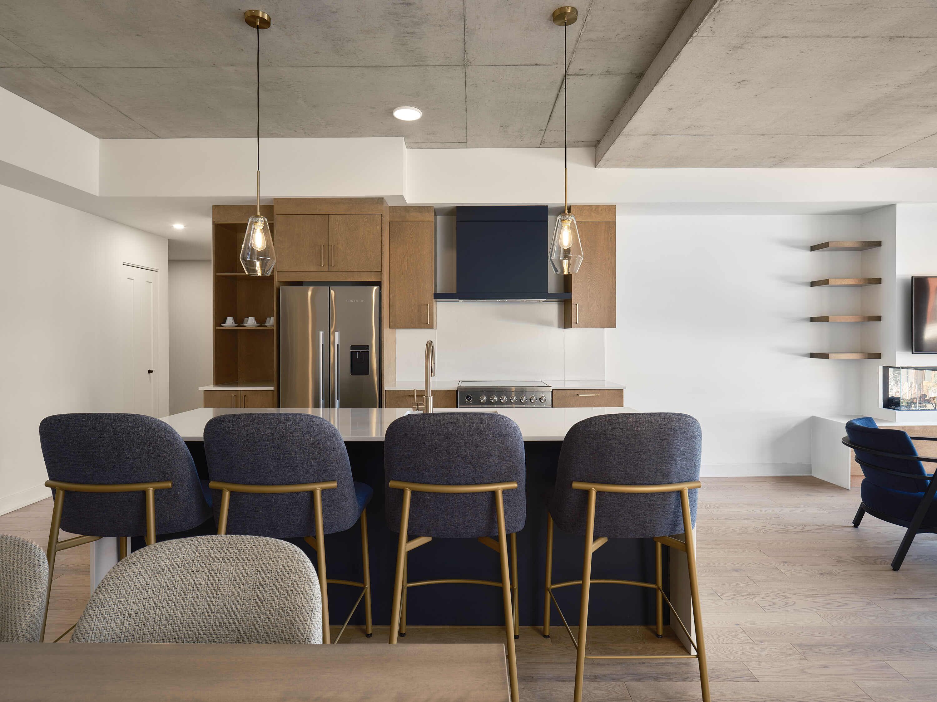 A modern kitchen with a stainless steel refrigerator, a built-in oven, and a dark blue backsplash. There are four bar stools with wooden legs and grey upholstery.