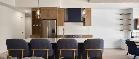 A modern kitchen with a stainless steel refrigerator, a built-in oven, and a dark blue backsplash. There are four bar stools with wooden legs and grey upholstery.