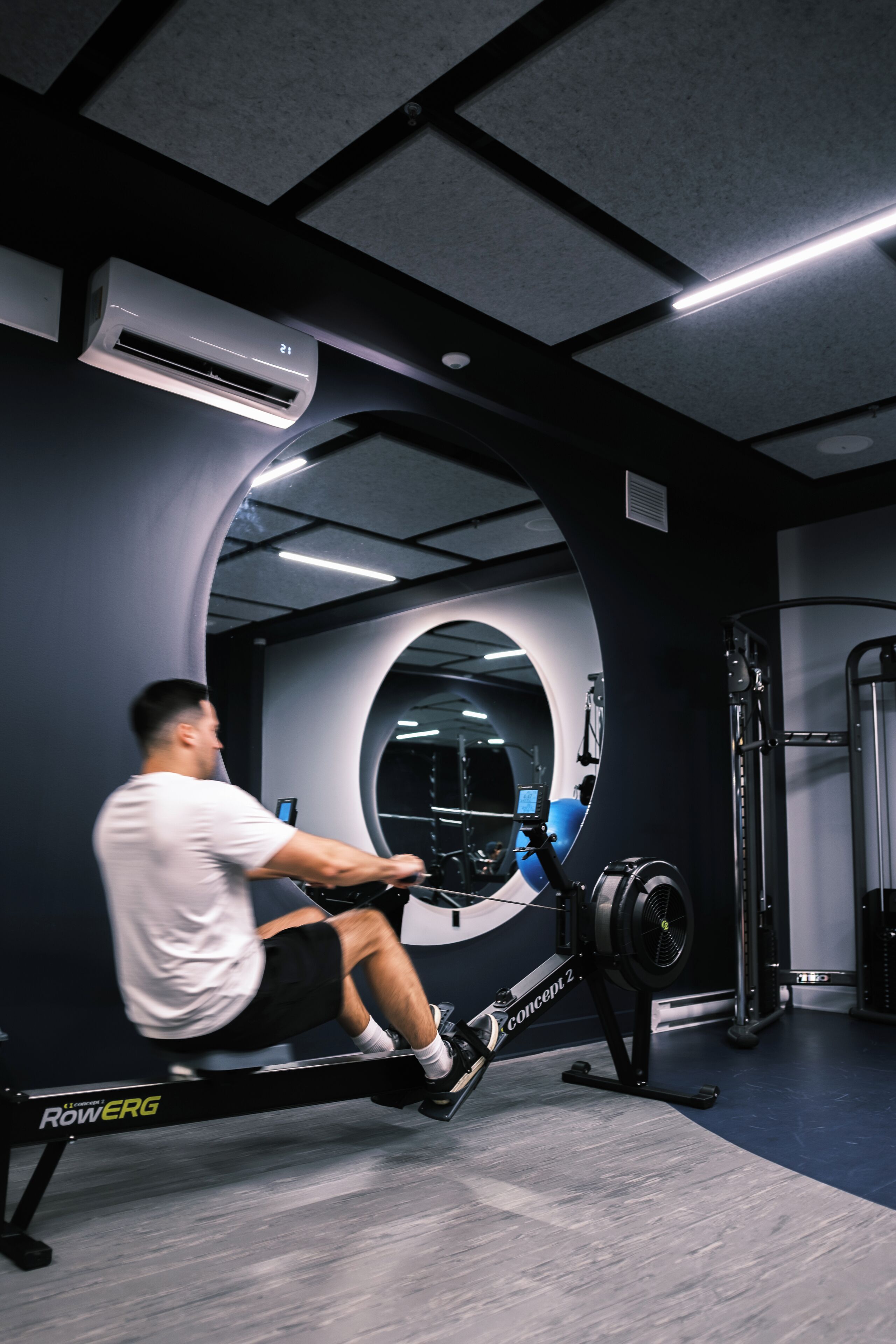 Een man gebruikt een roeimachine in een sportschool met een spiegelwand en een plafondventilator.