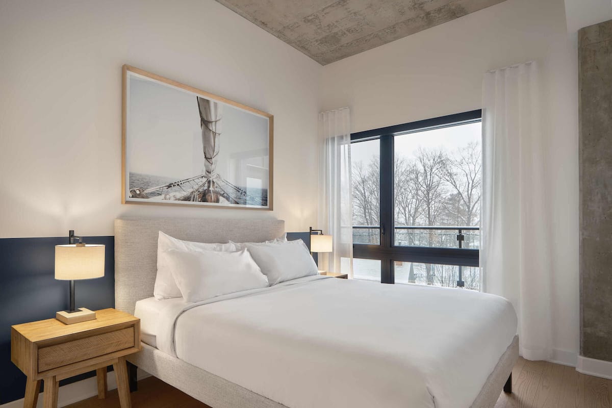 A hotel room with a large bed, two bedside tables with lamps, a framed picture above the bed, and a large window with a view of trees.