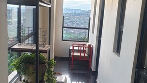 Ein Korridor mit Blick auf eine Stadtlandschaft durch ein großes Fenster, Regale mit Blumentöpfen und ein roter Sessel.