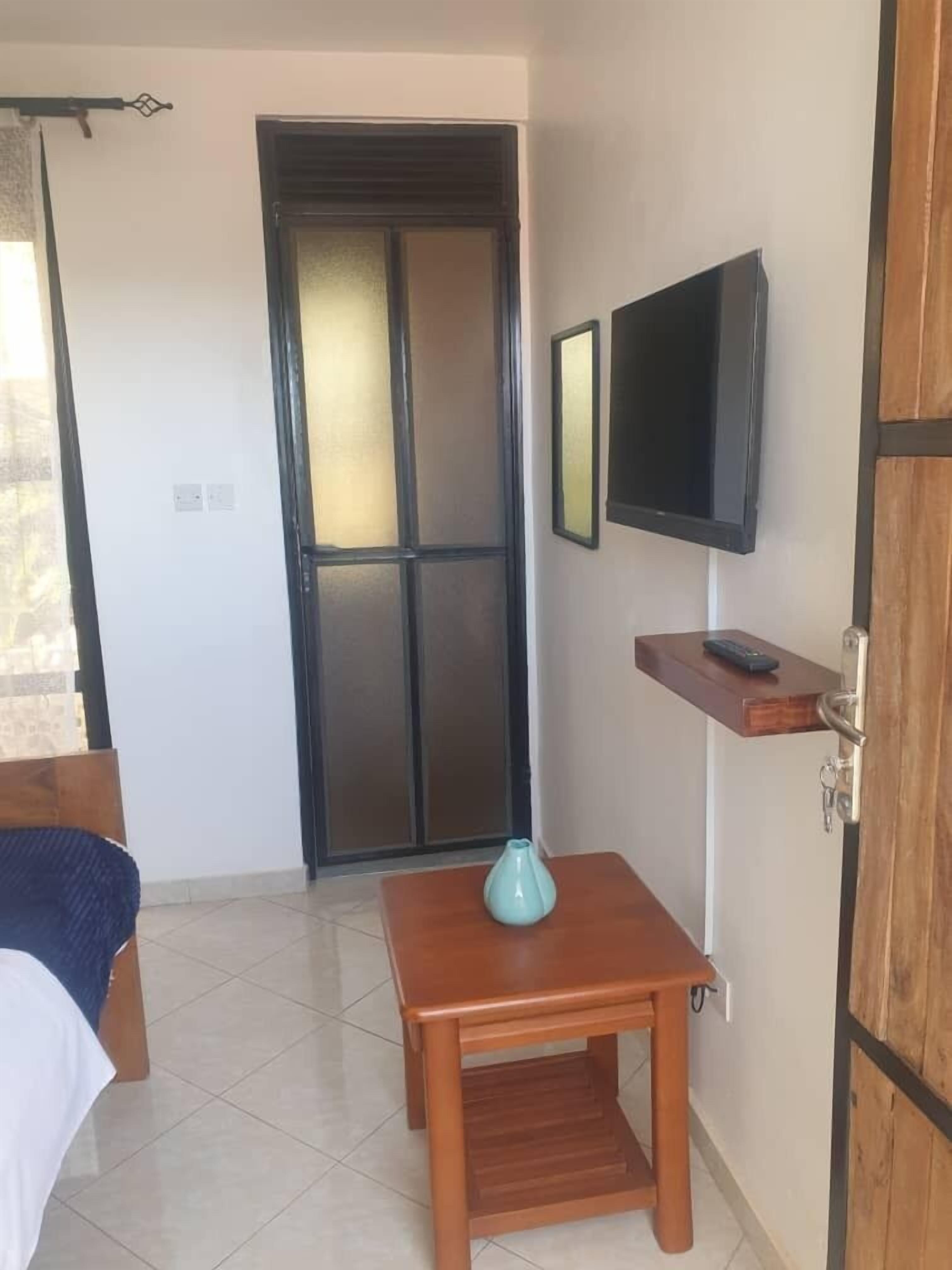 Habitación de hotel con cama, una pequeña mesa de madera con un jarrón, televisor montado en la pared y una puerta con panel de vidrio corredera.