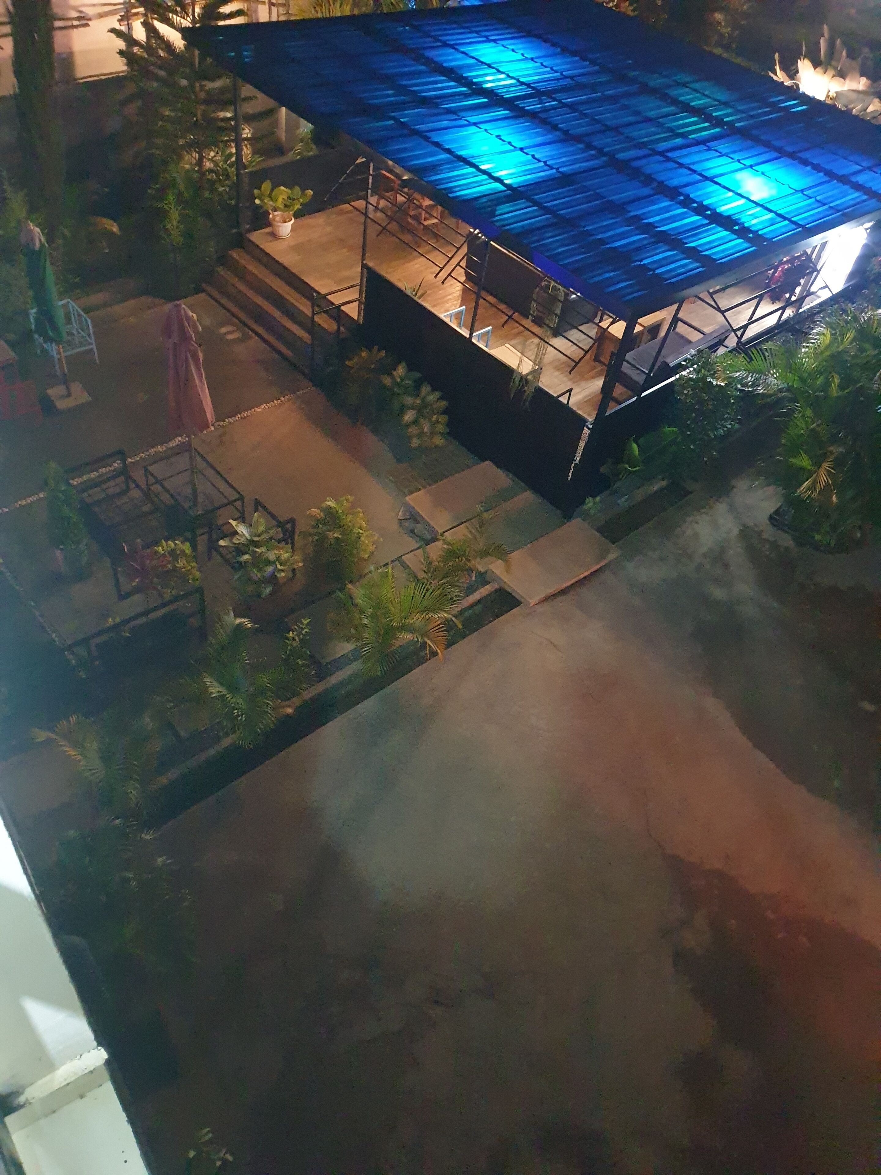 An aerial view of a rooftop with a blue-tinted structure, surrounded by greenery and a paved area.