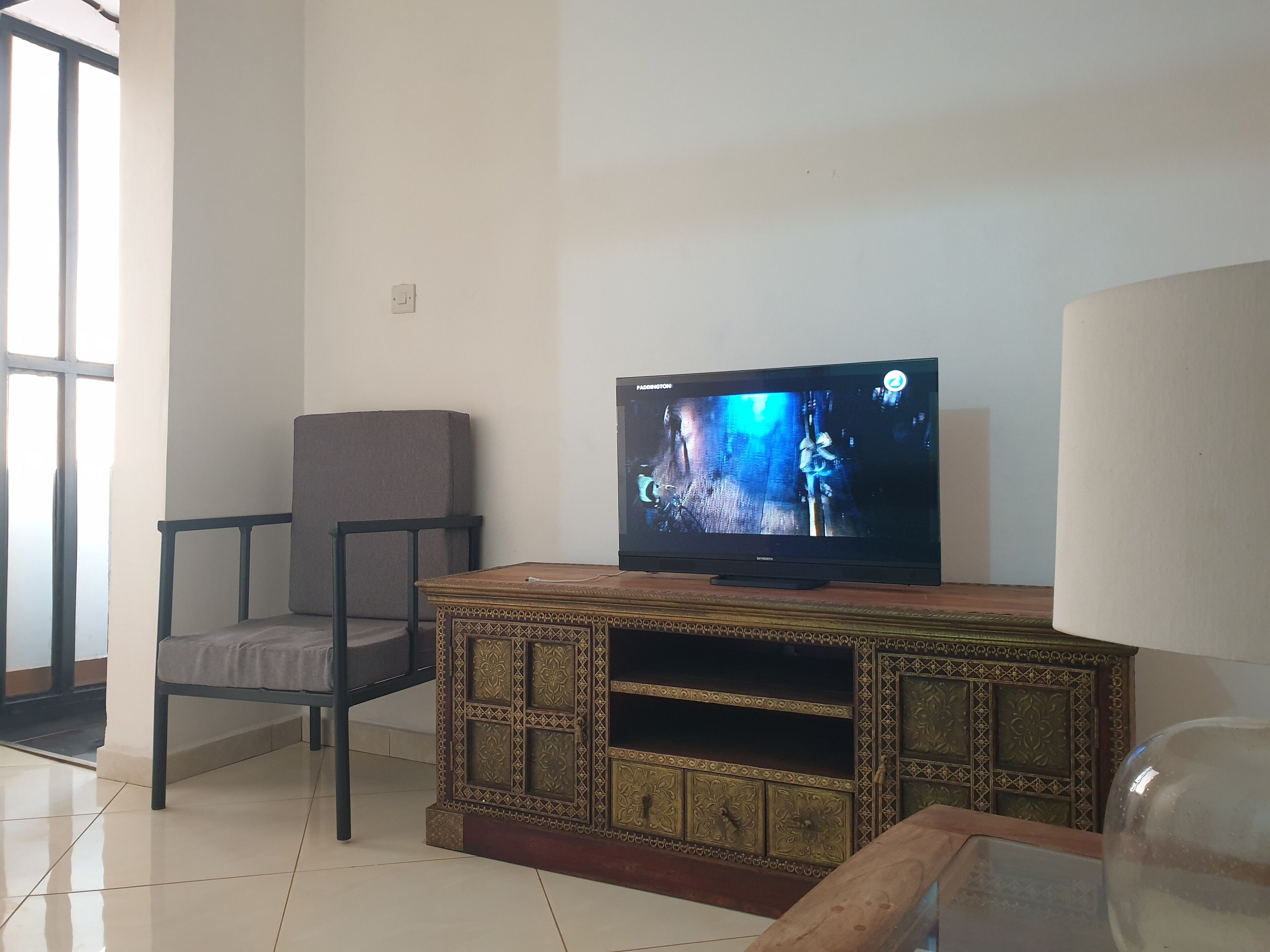 A living room with a television on a wooden cabinet, a grey chair, and a glass coffee table.