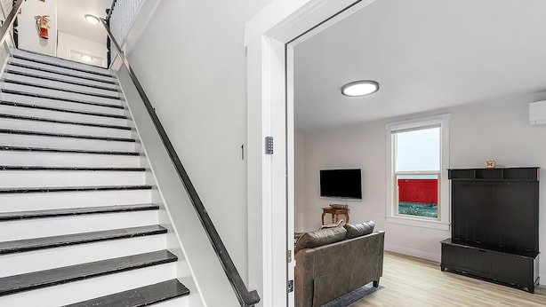 A staircase leading to a living area with a sofa, a flat-screen TV, and a window with a red view. - The Nighthawk Eyrie (Stanwood)