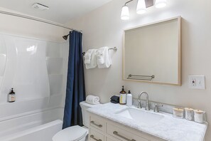 A bathroom with a bathtub, shower, toilet, and a sink with toiletries. - The Nighthawk Eyrie (Stanwood)