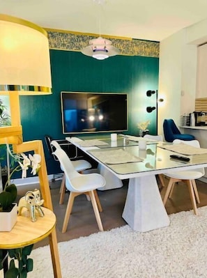 Ein modernes Wohnzimmer mit einem Glastisch, weißen Stühlen, einem großen Flachbildfernseher und einer wandfarblich tealfarbenen Akzentwand.