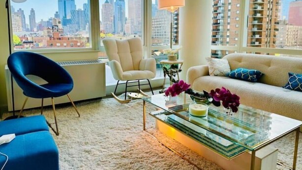 A modern living room with a city view, a blue chair, a beige sofa, and a glass coffee table. - Times Square Luxurious Massive 2bed/2bath flat,NYC (New York)