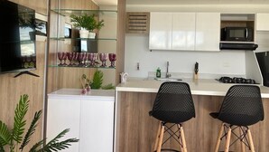 A modern kitchen with white cabinets, a black microwave, and a gas stove. There are two black chairs and a glass shelf with plants and decorative items. - Edificio Avenida Malecon (Guatapé)